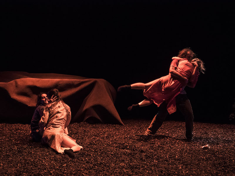 One couple sits on the peet-covered floor. One dancer touches their hand to the other's cheek. Another couple embrace one another. The women's legs swing in the air. A tent-looking shelter is in the background. 